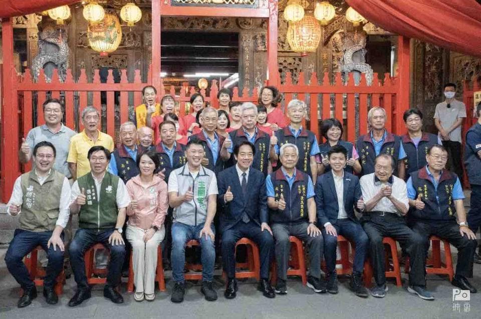 這張照片是一群人在廟宇前合照,他們都穿著深色外套,有些人還戴著帽子,表情看起來都很開心。