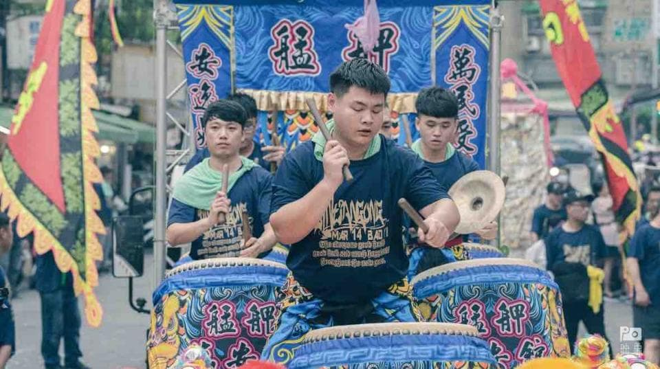 這張照片是三個年輕人正在敲擊大型的鼓,他們身穿藍色的衣服,站在一個裝飾華麗的平臺上。