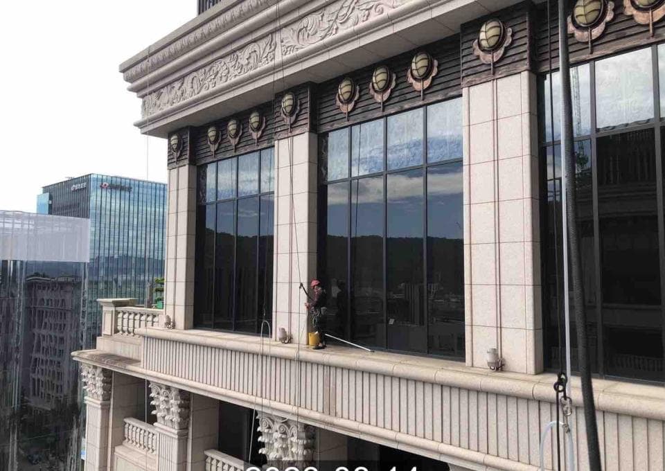1) 照片顯示一座高樓大廈的外觀,建築外牆是淺色石材,窗戶很大且乾淨,有一名身穿安全裝備的清潔工正在清洗窗戶
