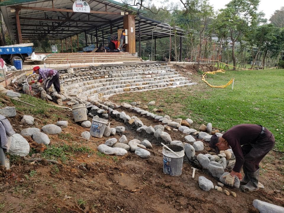 這張照片是兩位工人正在用石頭建造一個階梯。他們正在用水泥將石頭固定在一起,並且在階梯上鋪設一些大石頭。