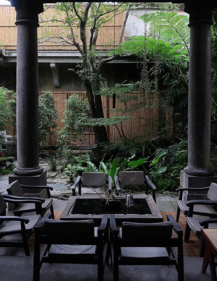 這張「庭院」的照片是,一個被綠色植物環繞的露臺,有幾張黑色的椅子和桌子,中間是一個方形水池。