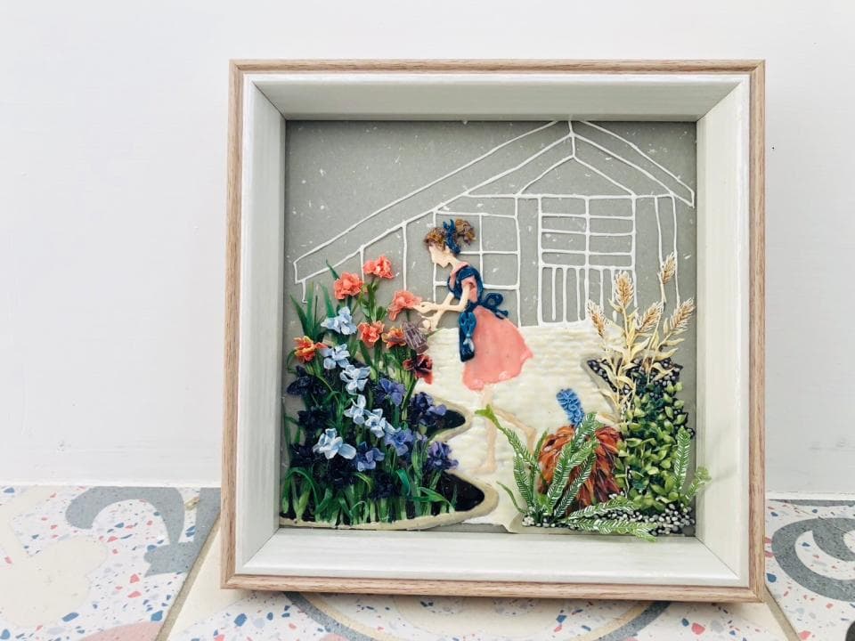 這張照片是一幅用手工製作的花朵和人物組成的立體畫,畫中一位身穿粉紅色連衣裙的女孩正在花園中採花。
