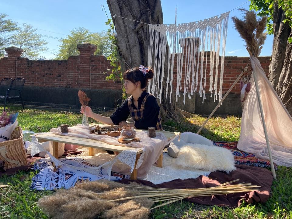 這張戶外野餐的照片是一個女生坐在野餐桌旁,桌子上擺著食物和飲料,旁邊有一個帳篷和一些裝飾品。