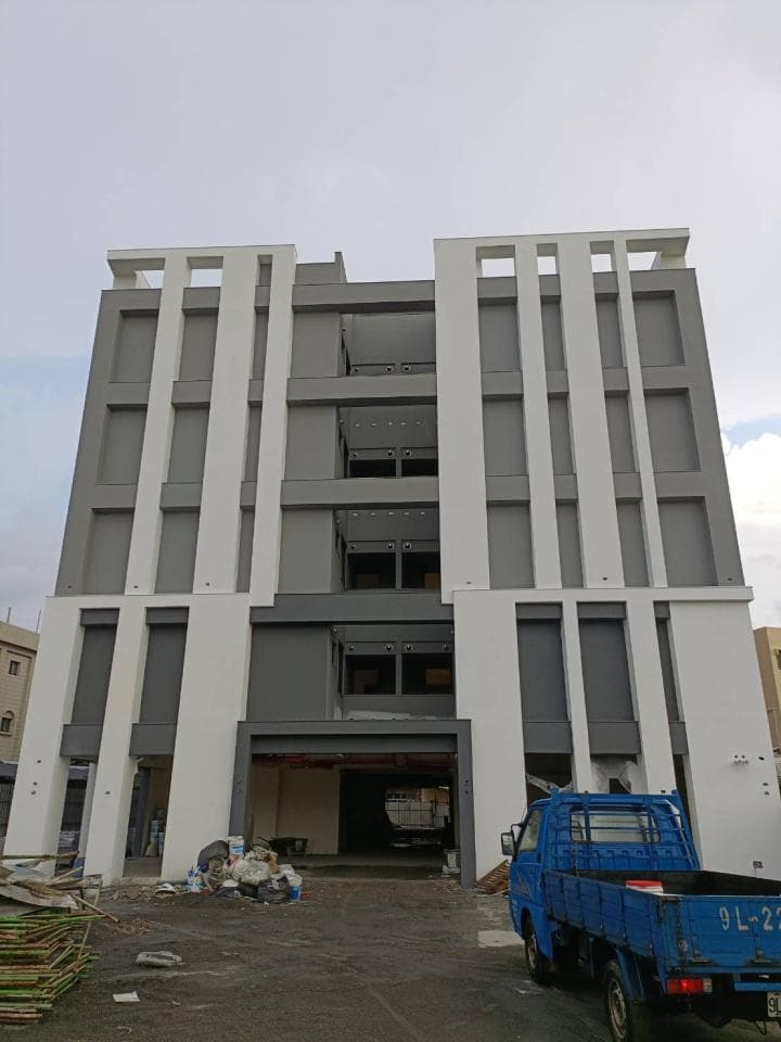 這張照片是一棟正在施工中的建築物,建築物上有許多窗户,窗户邊框是白色的,建築物的外牆是灰色的,照片中還有一輛藍色的貨車。