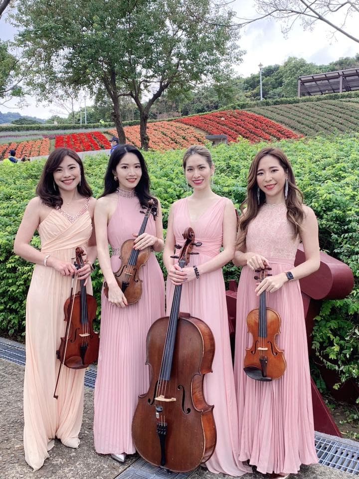 這張照片是四位穿著粉紅色禮服的女性,她們站在花園中演奏古典樂器。她們手中的樂器,分別是小提琴和大提琴。她們的動作優雅,音樂聲似乎在花園中迴盪,呈現出一幅美好的景象。