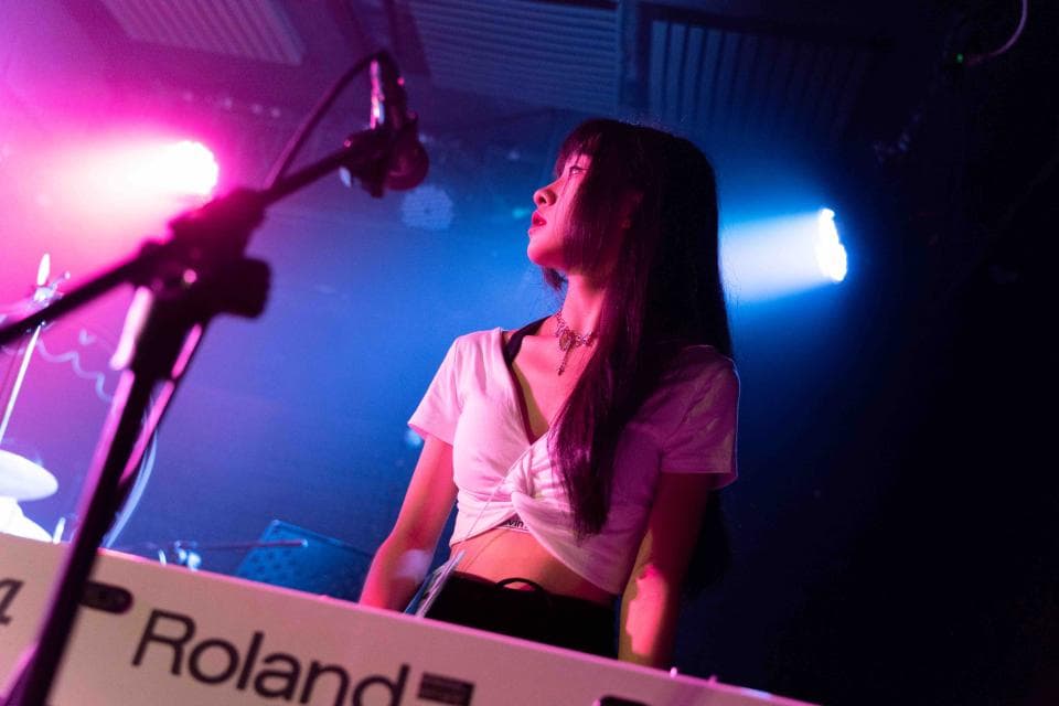 這張照片是一位年輕女性在舞臺上彈奏鍵盤。她身穿白色上衣,黑色褲子,頭戴耳機,正在專注地演奏。舞臺上還有麥克風架和燈光,營造出熱鬧的表演氛圍。