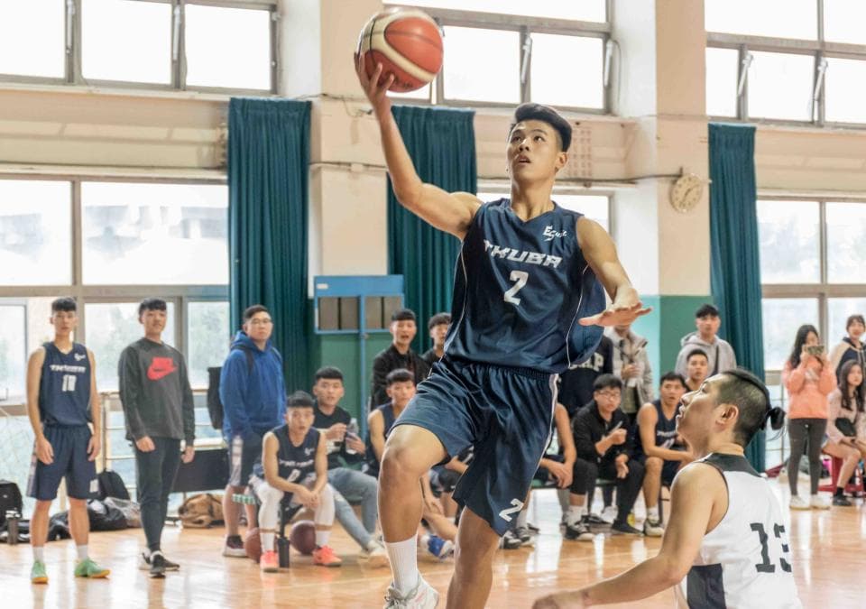 這張照片是一個籃球比賽,照片中最重要的部分是正在投籃的球員。他穿著藍色球衣,正在籃球架下投籃,表情專注,動作流暢。他身旁還有許多觀賽的球員,他們都注視著他。