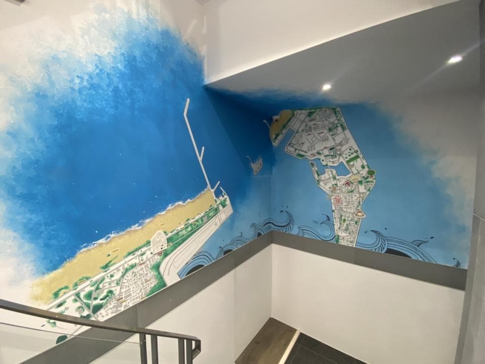 這張照片是一面牆壁上的壁畫,描繪了藍色的海洋和海岸線。最顯眼的是右邊的島嶼地圖,上面有綠色的草地和街道,細節豐富。