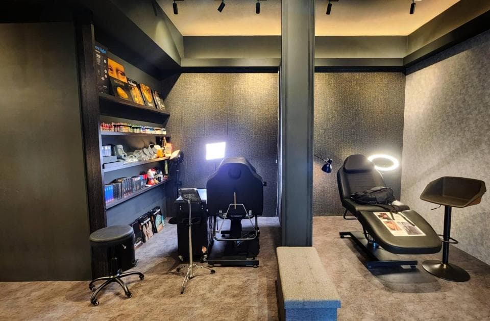 這張「美容工作室」的照片是一個現代化的空間,帶有黑色和灰色調。照片中可以看到一個黑色的美容床,一個黑色的凳子,一個黑色和銀色的置物架,以及一些美容設備。