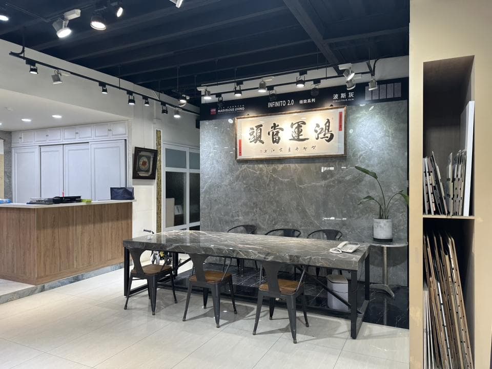 這張「家居裝修」的照片是一個現代化的展廳,展廳的牆上有一塊大理石牆,牆上掛著一個中國書法牌匾,展廳裡擺放著一張黑色的長方形桌子和幾把黑色的金屬椅子。