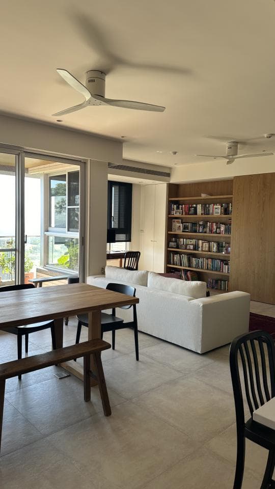 這張照片是一間現代化客廳,有一個木質餐桌和長椅,以及一個大型白色沙發。餐桌旁邊有一個白色風扇,為房間增添了一絲涼意。