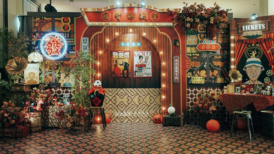 這張照片是一個復古風格的裝飾,有紅色的燈籠、老式的霓虹燈招牌和木製的裝飾。舞臺背景是紅色的幕布和金色的裝飾,還有幾張桌子和椅子。