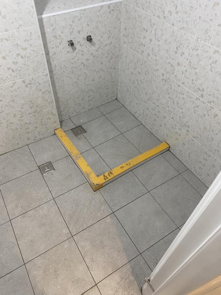 這張「浴室」的照片是裝修完的浴室,地板是淺灰色的瓷磚,牆壁上是淺色花紋的瓷磚,照片右邊可以看到一塊白牆,照片底部有一塊黃色木條。