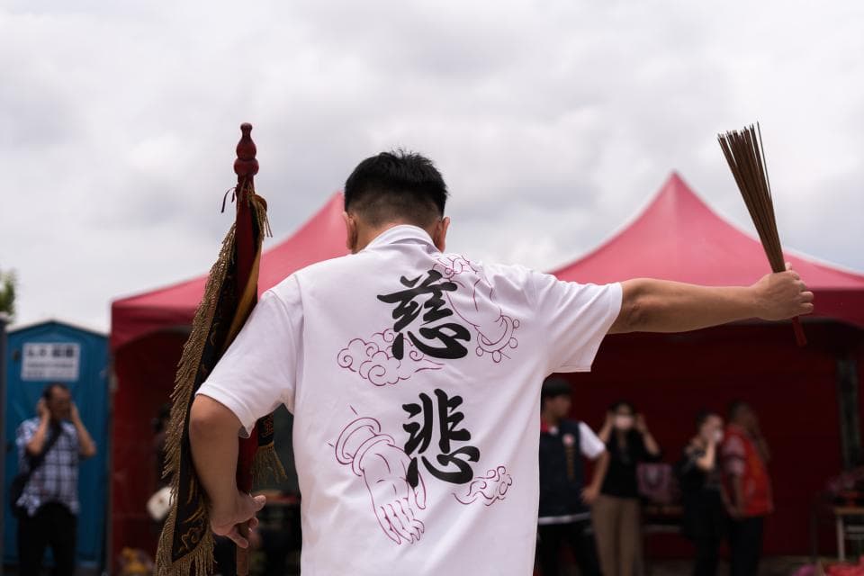 這張照片是一個男子在儀式中舉著香,他穿著印有漢字的白色襯衫,襯衫上的字看起來像是「慈悲」。男子身後是一些紅色的帳篷,畫面充滿宗教儀式感。