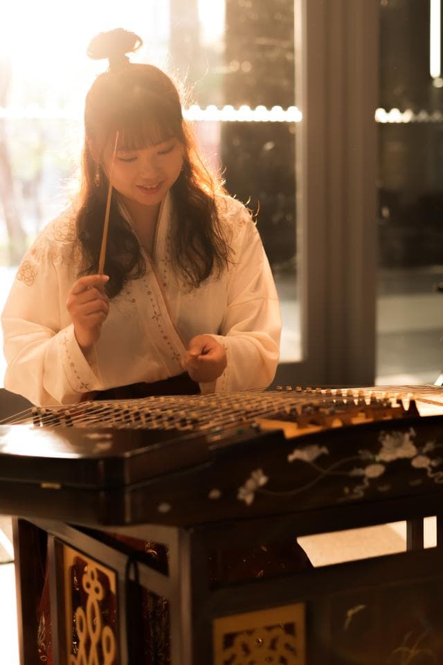 這張「一位女子在彈奏古箏」的照片是,女子穿著白色古裝,神情專注地彈奏著古箏,古箏上雕刻著精美的圖案,背景是窗戶和光線,營造出古典優雅的氛圍。