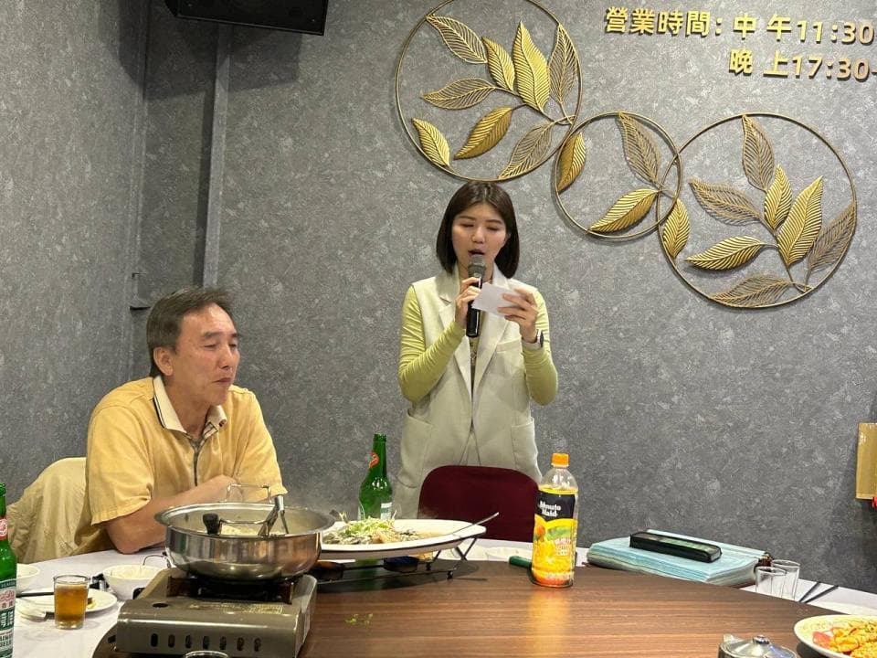 這張「餐廳」的照片是一個正在舉行活動的餐廳,照片中有一位女士正在臺上講話,旁邊有一位男士在臺下聆聽,桌上放著一些食物和飲料。