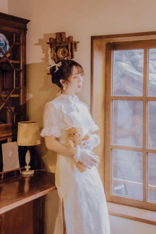這張照片是一位穿著白色蕾絲連衣裙的年輕女性,她站在窗邊,抱著一隻毛絨玩具。房間裡有一個木製的書架和一張桌子。