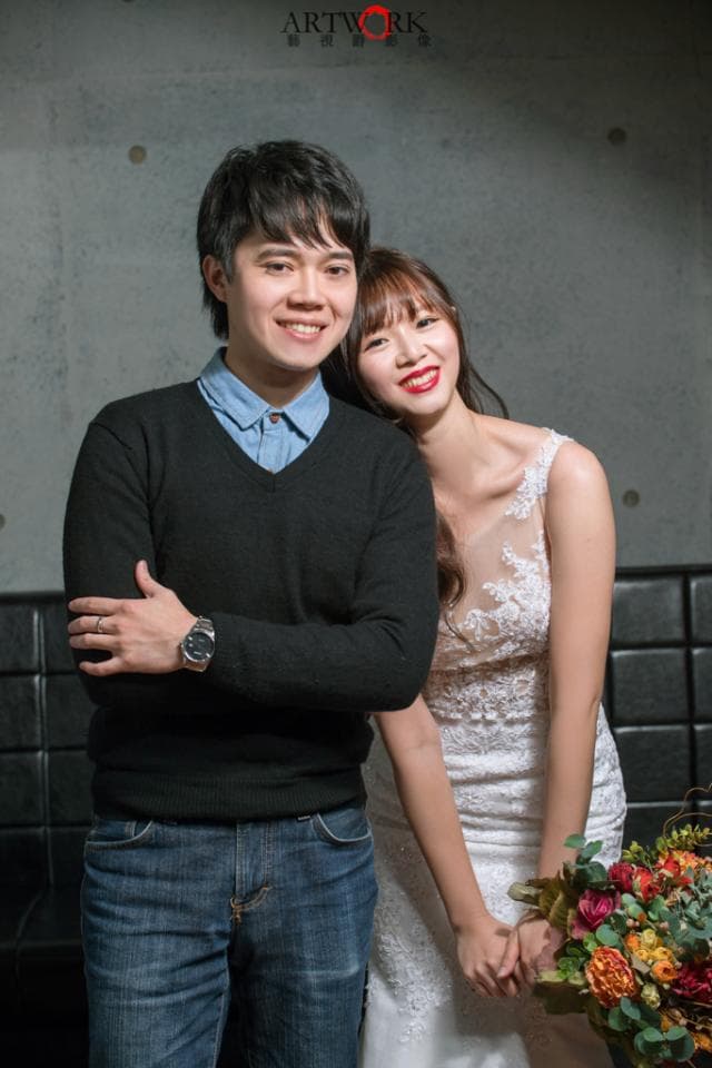 這張照片是一對男女的婚紗照,他們站在水泥牆前,彼此相依偎,眼神充滿著愛意。男方穿著黑色毛衣和牛仔褲,女方則穿著白色婚紗,手裡拿著鮮花,整體畫面充滿甜蜜和幸福。