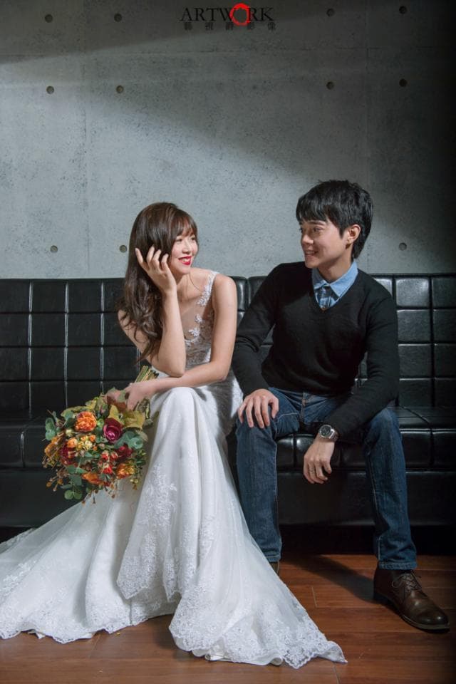這張「婚紗照」的照片是,一對新人在黑色沙發上,新娘穿著白色婚紗,新郎穿著黑色上衣和牛仔褲,他們互相看著對方,新娘手裡拿著一束花。