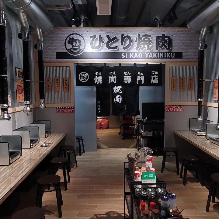 這張「燒肉店」的照片是一個日式燒肉餐廳,餐廳內部有許多座位和抽油煙機,以及一個櫃臺。牆上掛著日文招牌,上面寫著「ひとりの焼肉」,意思是「一人燒肉」。