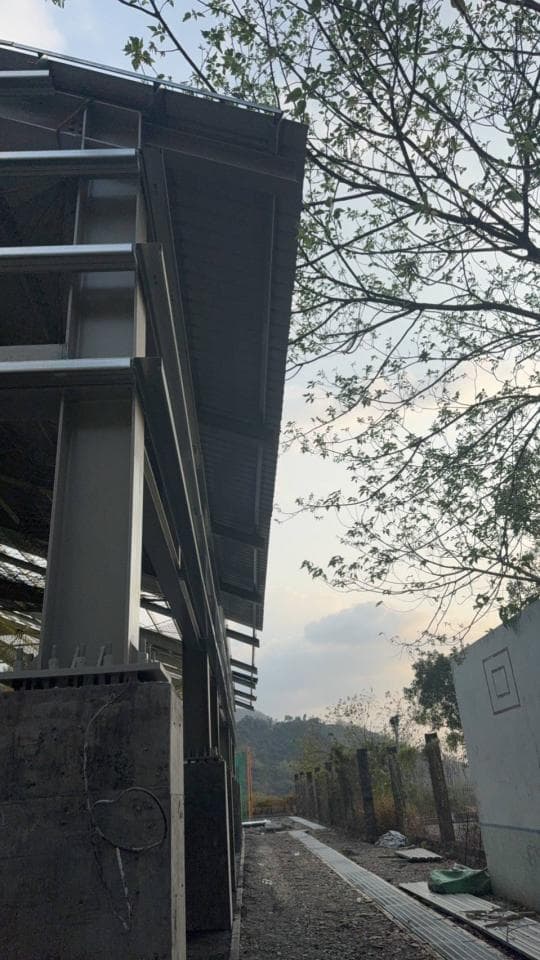 這張建築照片是一個鋼結構建築,可以看見建築的鋼骨架和部分屋頂。建築外觀看起來很新,旁邊有一棵樹和一些綠色植物。