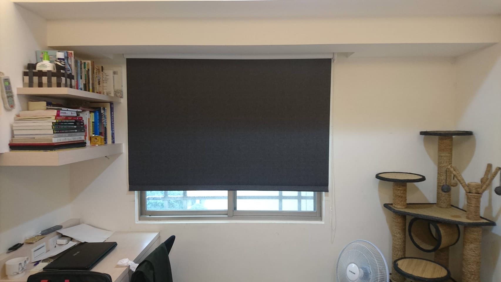 1) 照片顯示一個房間的一角,房間裡有一扇窗戶,窗戶上裝有窗簾,窗簾是灰色的,窗戶下放著一台筆記型電腦,房間牆壁上有一個層架,層架上有許多書本,房間牆壁上還有一個貓咪跳台