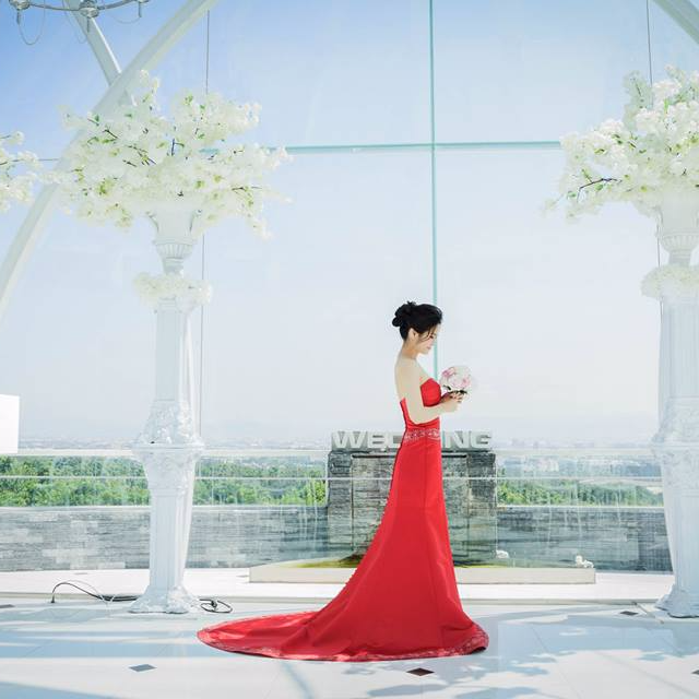 這張照片是一個穿著紅色婚紗的女性,她站在一個玻璃建築物中,旁邊有兩盆白色的花。