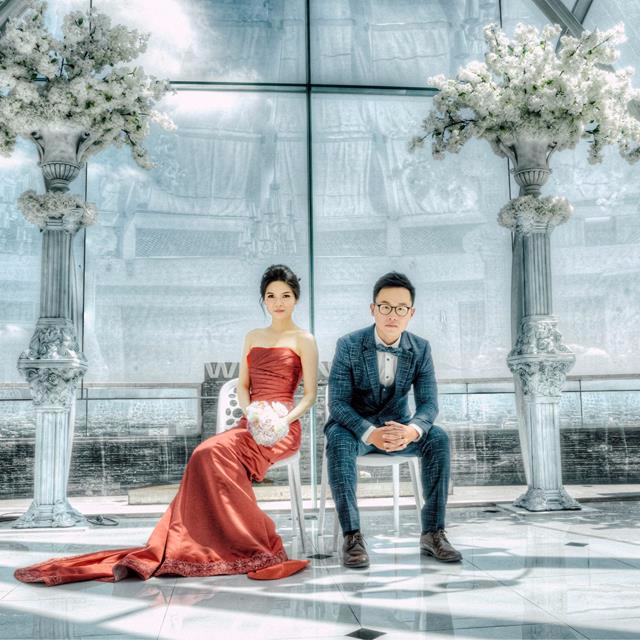 這張照片是一對穿著婚禮服裝的男女,他們坐在白色椅子上,背景是一片透明的窗戶,窗戶外有兩棵白色花樹。他們看起來非常幸福,準備好迎接他們人生的新篇章。