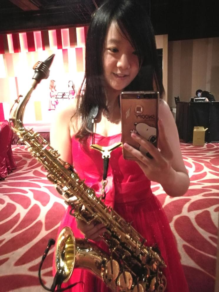 這張照片是一個年輕女子手持薩克斯風的肖像照。她穿著紅色連衣裙,並微笑著看向鏡頭。她的手指放在薩克斯風的按鍵上,暗示她是一位音樂家。
