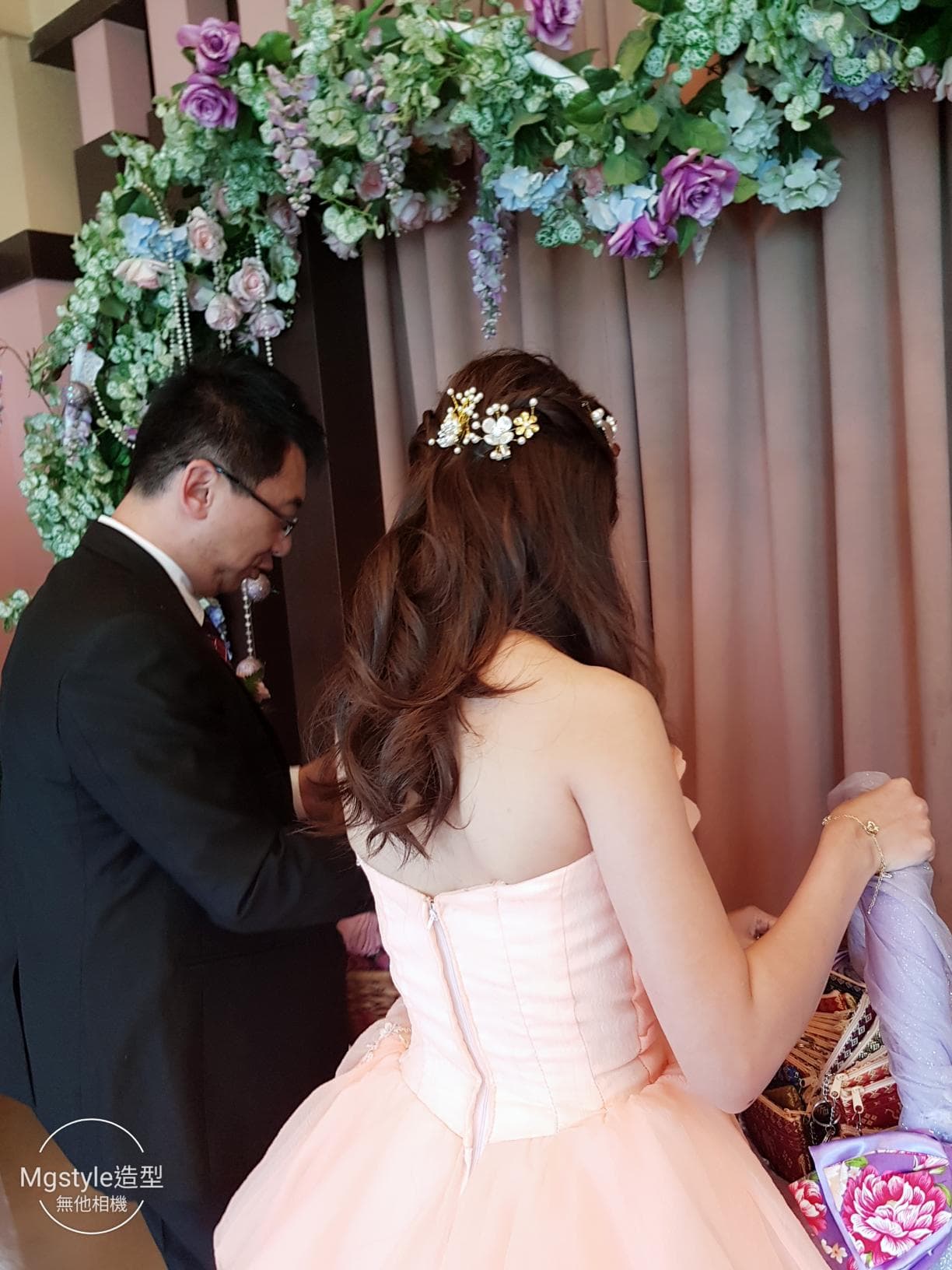 這張婚禮的照片是一對新人在一個佈置著鮮花的拱門下合影,新娘穿著一件粉色的婚紗,新郎穿著一件黑色的西裝。