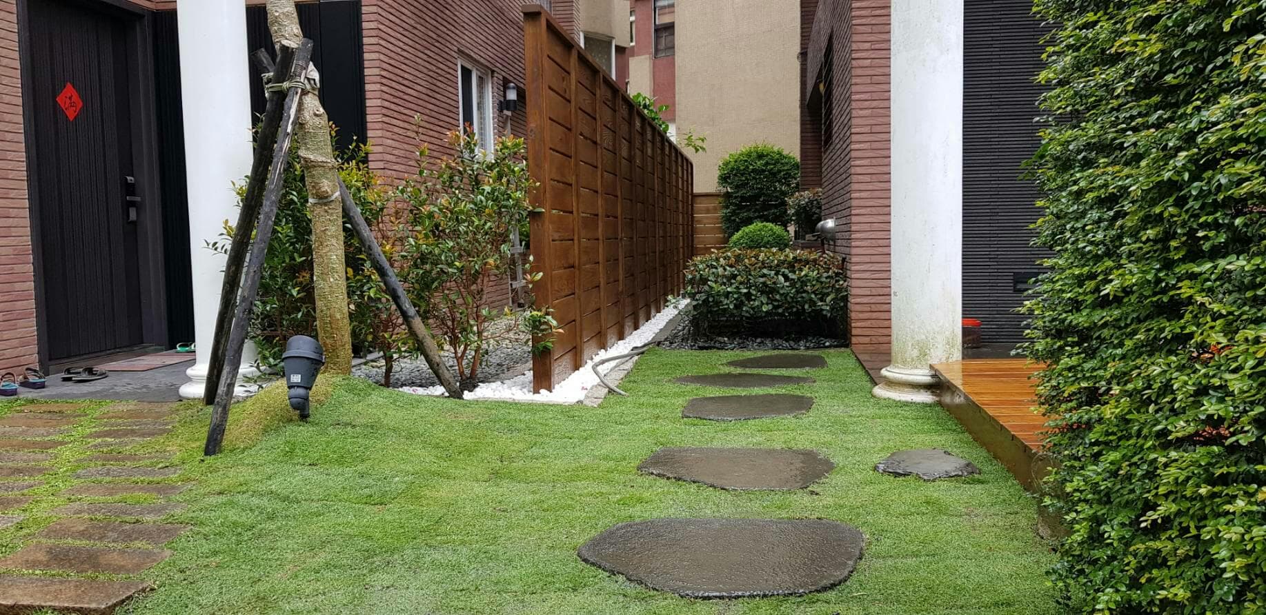 這張照片是 一個有石頭路徑、綠色草坪、木頭圍籬和磚牆的小花園。