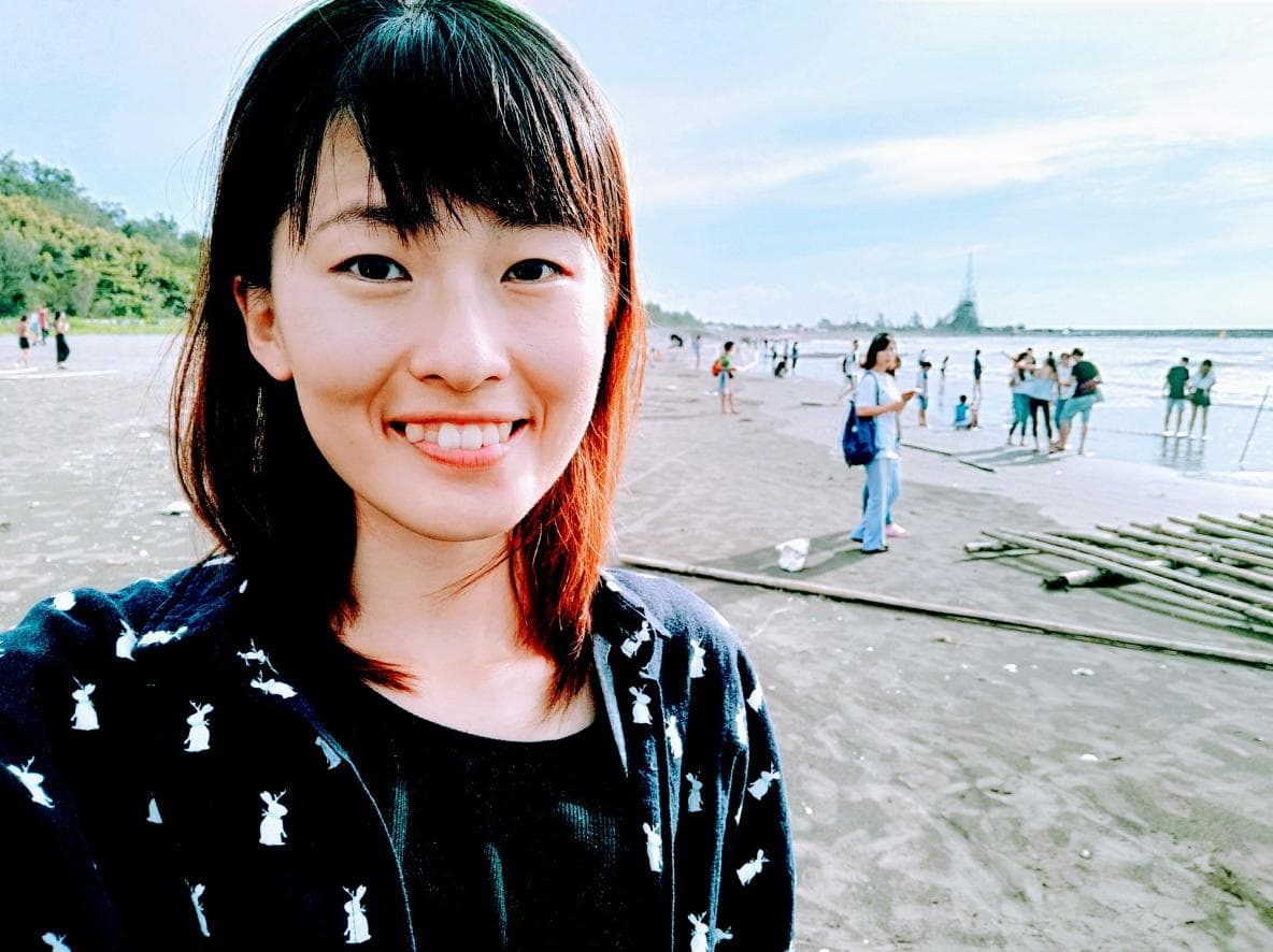 這張照片是一個年輕女性站在海灘上的自拍照,她穿著一件黑色外套,背景是沙灘和一些遊客。