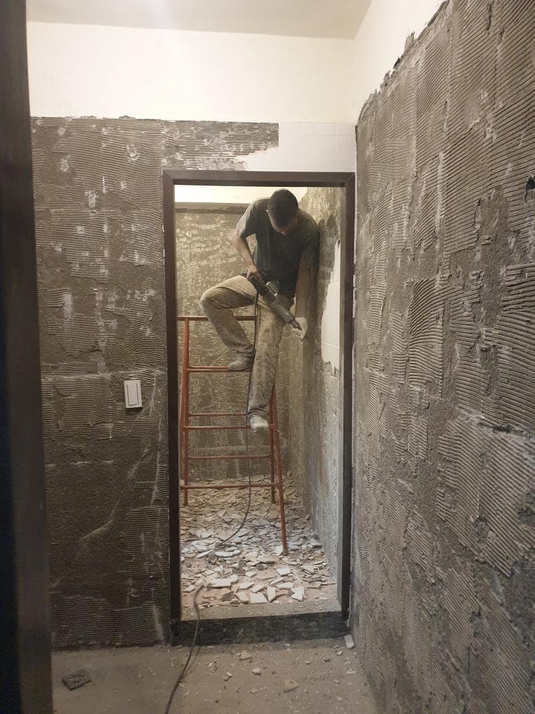 一名男子站在梯子上,使用電鑽敲打牆壁,牆壁上布滿了水泥塊和灰塵,周圍環境看起來是正在進行房屋整修