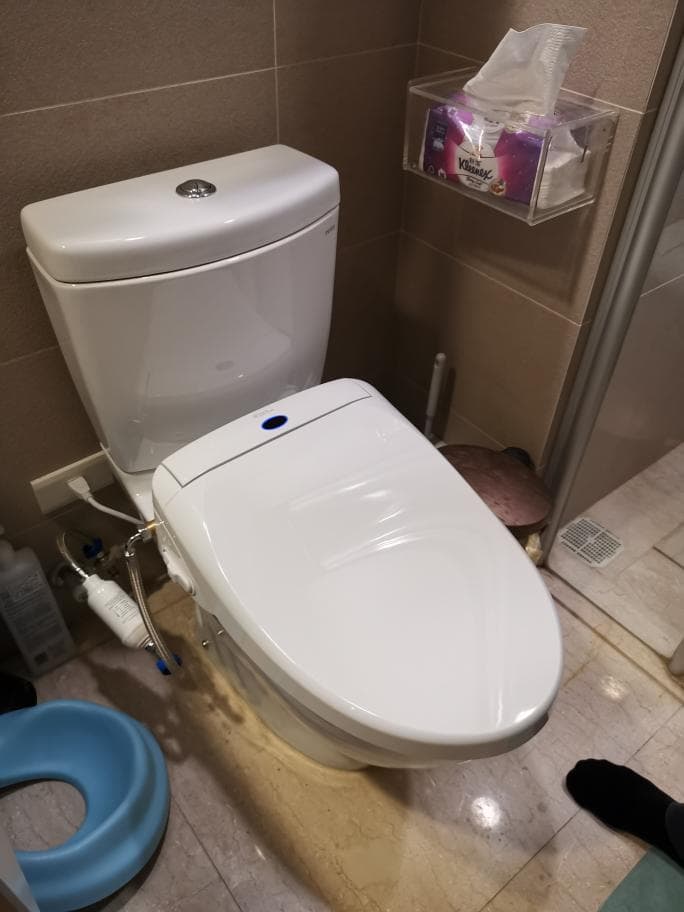 1) 照片顯示一個白色馬桶,馬桶蓋上有一個藍色按鈕,馬桶上有一個水箱,看起來是新的,地板上有一個藍色的兒童馬桶,馬桶旁邊有一個組織盒,組織盒是透明的,裡面有粉紅色的衛生紙,馬桶附近還有水管和一個水壺