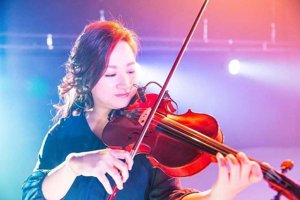 1) 照片中一位年輕女性身穿藍色上衣,手持小提琴演奏,舞台背景為紫色和紅色燈光