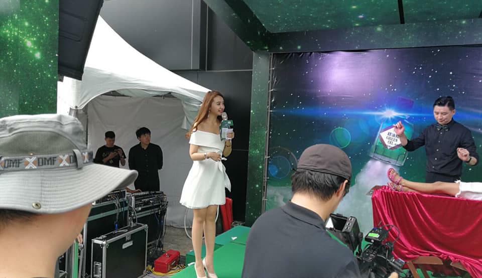 這張照片是一場魔術表演,舞臺上有一位穿著白色禮服的女主持人,一名男魔術師正在表演魔術,一位女性觀眾躺在地上,似乎被男魔術師施了魔法。舞臺前有幾個人在觀看錶演。