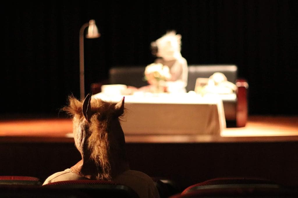 這張「劇場演出」的照片是一個戴著動物頭套的人坐在觀眾席上,舞臺上有一個模糊的人影,背景是一盞燈光。