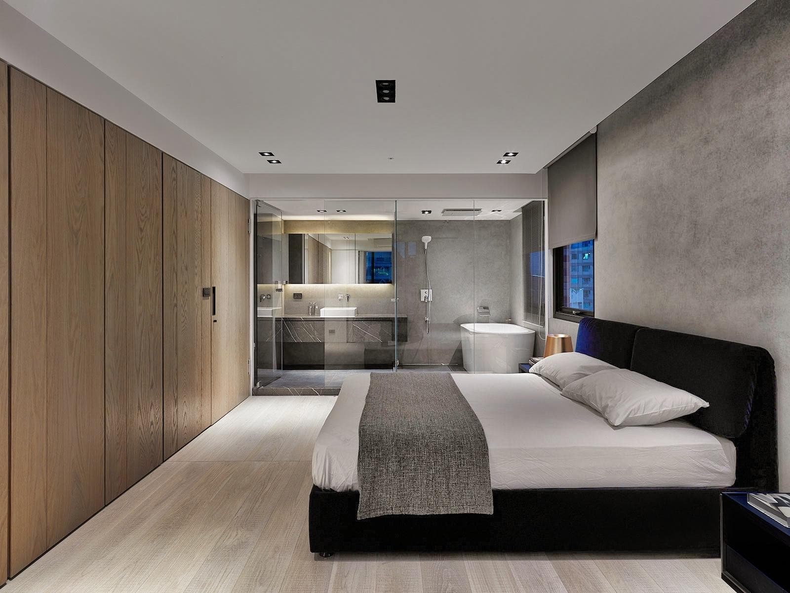 這張臥室的照片是一個現代化的臥室,有著木質的牆面和玻璃的浴室,床頭板是黑色的,床單是白色的,床頭櫃上放著一個黑色的檯燈。