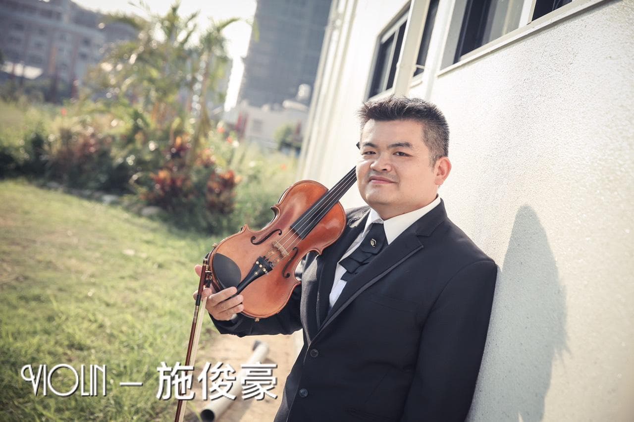 1) 照片中是一位身穿黑色西装的年輕男子,他站在一面白色牆壁前,手里拿着一把小提琴