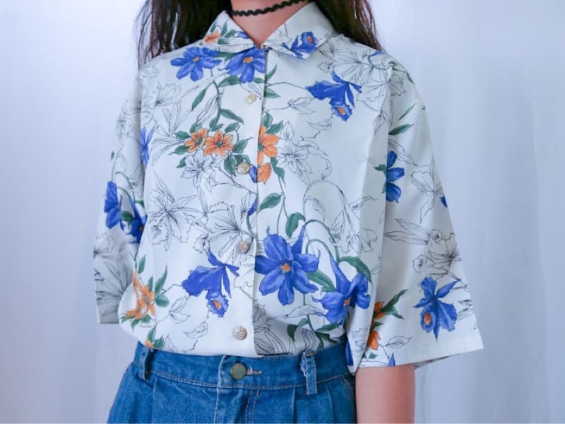 這張「白色襯衫」的照片是,襯衫上有藍色、橙色和綠色的花卉圖案。襯衫是短袖的,領口是翻領,而且有金色的鈕扣。襯衫搭配了一條牛仔褲。