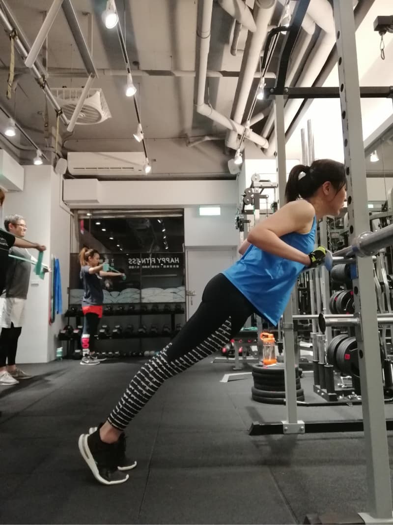 這張照片是一個女人在健身房做俯臥撐。她穿著藍色背心和黑白條紋運動褲。她握著健身器材,身體保持穩定。