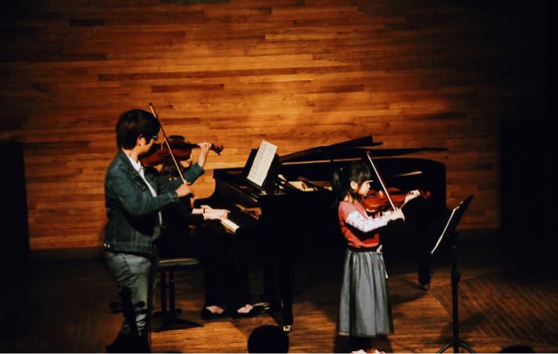 這張「音樂表演」的照片是兩個女生正在演奏小提琴,其中一位在演奏鋼琴,她們正在一個木頭地板的舞臺上表演。