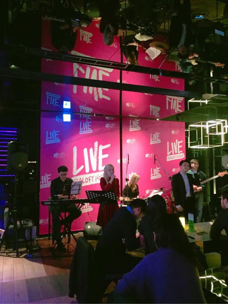 這張照片是現場演奏的場景,中間是一位穿着紅色裙子的女歌手正在唱歌,旁邊的樂隊正在伴奏。