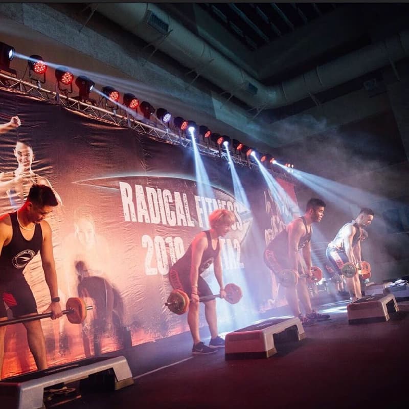 這張照片是四個人在舞臺上舉重,背景是一個寫著「RADICAL FITNESS 2019」的佈景。他們正在參加一個健身比賽。