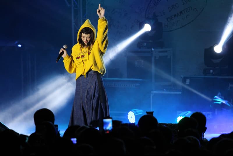 這張「演唱會」的照片是一個女生歌手在臺上唱歌,她穿著黃色的外套和藍色的裙子,正在用麥克風唱歌,臺下有很多觀眾正在觀看演出。