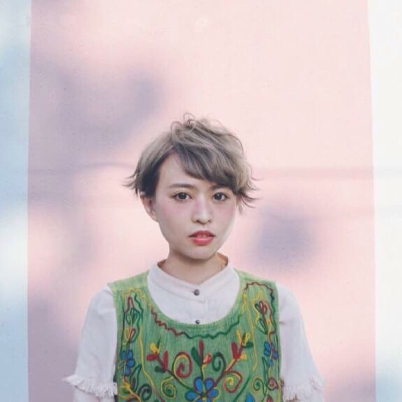 這張照片是一個年輕女子,她穿著一件綠色繡花背心和一件白色襯衫,她的頭髮是短的灰棕色,並且她正在微笑。