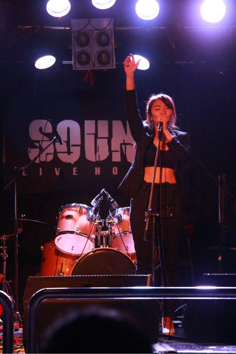 這張「現場演唱」的照片是一個女歌手在舞臺上唱歌,她穿著黑色西裝外套和黑色褲子,拿著麥克風,背景有鼓組和音響設備。