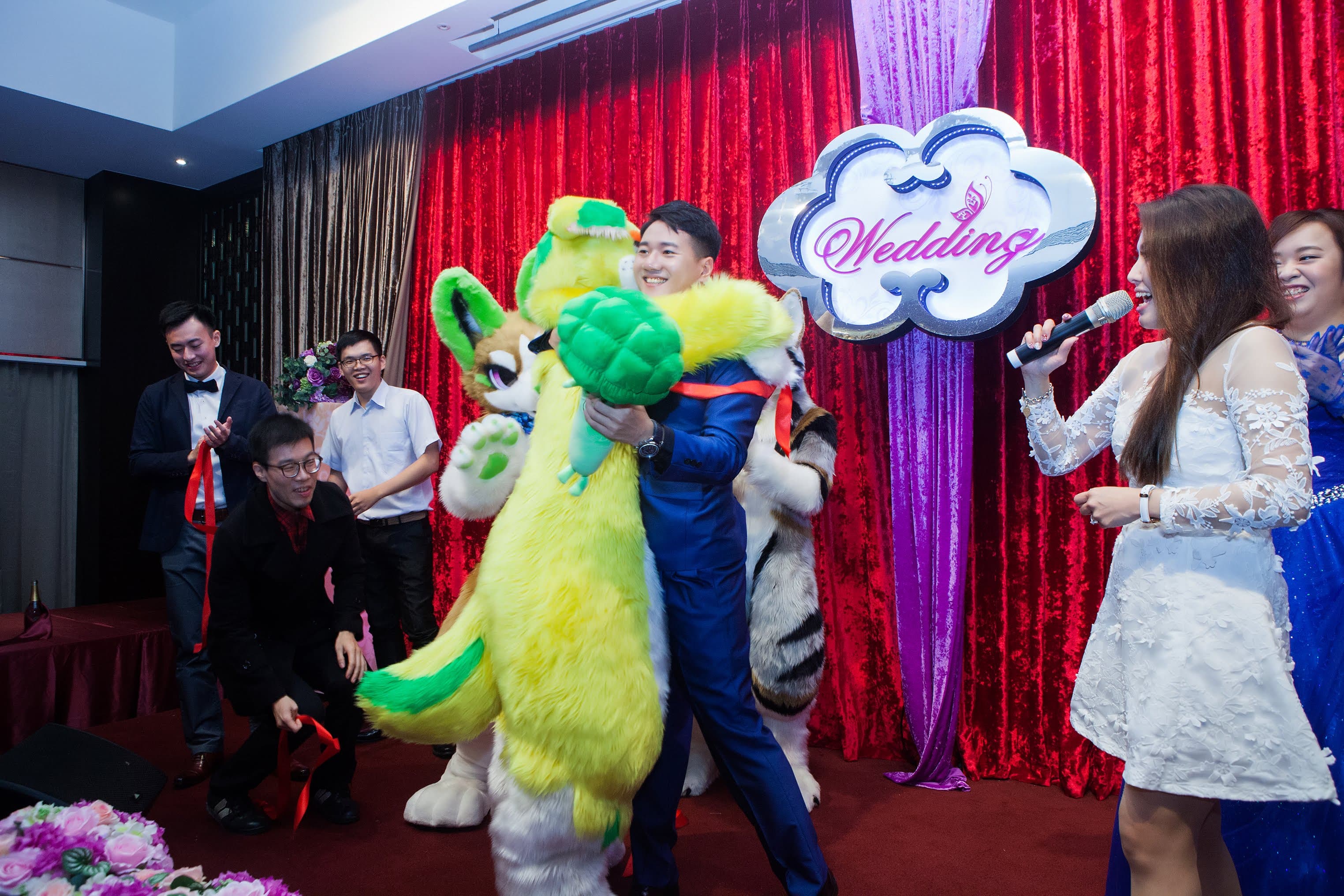 這張「婚禮」的照片是一對新人,新郎穿著西裝,新娘穿著白紗,他們正在和一隻可愛的毛絨玩偶合影。旁邊還有一位女士,她穿著白色的禮服,正在唱歌。
