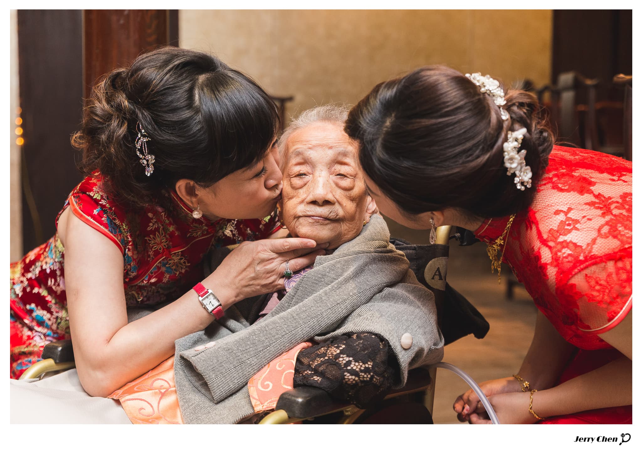 這張照片是一個老奶奶被兩個年輕女子親吻。老奶奶坐在椅子上,表情慈祥,而兩個女子都穿著紅色禮服,正在親吻老奶奶的臉頰。她們的動作顯得溫馨而甜蜜,展現了對老奶奶的愛與敬意。