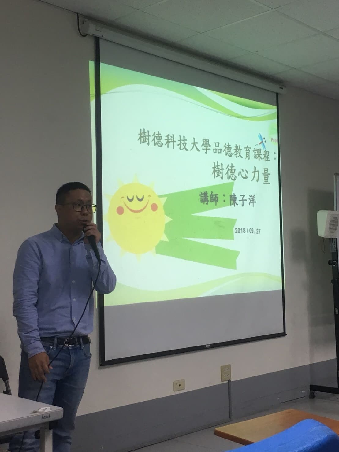 這張照片是一名男子在投影機前演講,投影機上顯示著「樹德科技大學品德教育課程:樹德心力量」。